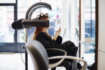 Mulher sentada sob secador de texto com telefone celular no salão de cabeleireiro — Fotografia de Stock