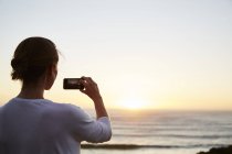 Жінка фотографує захід сонця над океаном з телефоном — стокове фото