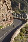 Male triathlete cyclist cycling uphill along sunny cliffs — Stock Photo