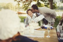 Boyfriend kissing girlfriend with birthday gift at lakeside patio table — Stock Photo