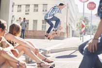 Друзі дивляться хлопчика-підлітка, який стрибає скейтборд на сонячному міському кутку — стокове фото