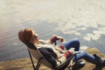 Serena mulher relaxante ouvir música com fones de ouvido e telefone inteligente na ensolarada doca à beira do lago — Fotografia de Stock