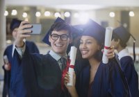 Выпускники колледжа в шапке и халате держат дипломы, позирующие для селфи — стоковое фото