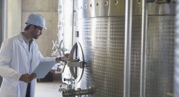 Vintner in lab coat and hard hat examining stainless steel vat in cellar — Stock Photo