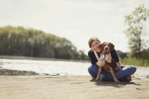 Retrato sorridente mulher abraçando cão na ensolarada doca à beira do lago — Fotografia de Stock