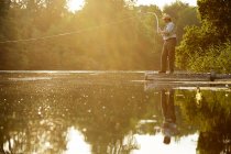 Uomo anziano pesca a mosca sul molo del lago estivo — Foto stock