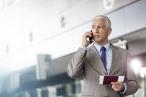 Businessman with passport and airplane ticket talking on cell phone in airport — Stock Photo