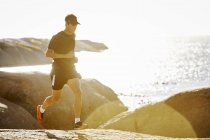 Triatleta masculino correndo em trilha rochosa ao longo do oceano ensolarado — Fotografia de Stock