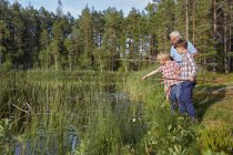 Grand-père enseignant petits-fils pêche au bord du lac ensoleillé — Photo de stock