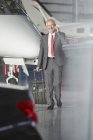 Усміхнений бізнесмен тягне валізу, розмовляючи на мобільному телефоні в ангарі літака — стокове фото