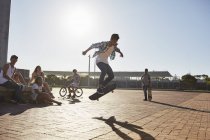 Amigos viendo adolescente volteando monopatín en el soleado parque de skate - foto de stock