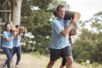 Determined man running with log on boot camp obstacle course — Stock Photo