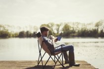 Hombre beber café y el uso de tableta digital en el muelle soleado junto al lago - foto de stock
