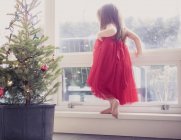 Girl in red dress on ledge next to potted Christmas tree — Stock Photo