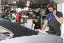 Mechanic talking on cell phone ordering car parts from catalog in auto repair shop — Stock Photo