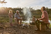 Grandparents and grandchildren enjoying campfire at sunny lakeside — Stock Photo