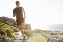 Мужской триатлонист бежит по солнечной скалистой тропе вдоль океана — стоковое фото