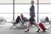 Empresaria tirando maleta más allá de hombre de negocios en reposo con almohada en el cuello en el área de salida del aeropuerto - foto de stock