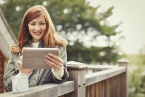 Усміхнена жінка з рудим волоссям використовує цифровий планшет і п'є каву на балконних перилах — стокове фото