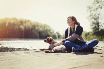 Mulher sorridente e cão relaxando na ensolarada doca à beira do lago — Fotografia de Stock