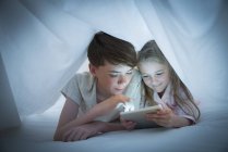 Брат и сестра делятся цифровыми планшетами под листом — стоковое фото