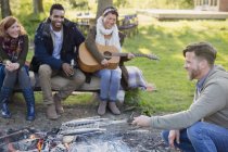 Friends playing guitar and cooking fish in grill basket over campfire — Stock Photo