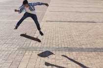 Teenage boy flipping skateboard on sunny cobblestone — Stock Photo