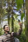 Sorrindo homem tomando selfie com câmera telefone em bosques ensolarados — Fotografia de Stock