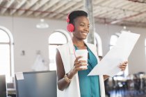 Smiling creative businesswoman with headphones and coffee reviewing paperwork in office — Stock Photo