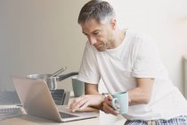 Homem de pijama bebendo café e trabalhando no laptop na cozinha — Fotografia de Stock
