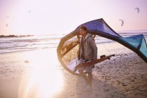 Uomo che trasporta attrezzatura kiteboarding sulla spiaggia al tramonto — Foto stock