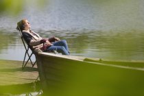 Mujer serena relajante escuchando música con auriculares en el muelle soleado junto al lago - foto de stock