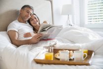 Couple souriant lecture journal profiter du petit déjeuner au lit — Photo de stock