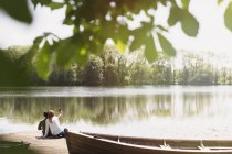 Casal com telefone de câmera tirando selfie na ensolarada doca à beira do lago ao lado da canoa — Fotografia de Stock