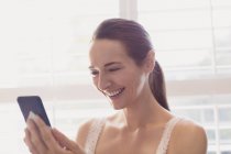 Mujer sonriente mensajes de texto con el teléfono celular en la ventana - foto de stock
