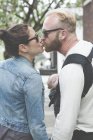 Vista de pareja en gafas de sol besando y sosteniendo al bebé en las calles de la ciudad - foto de stock