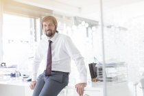 Retrato de homem de negócios sorridente vestindo camisa e gravata encostada na mesa no escritório — Fotografia de Stock