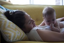 View of mother lying on sofa with little baby sucking thumb — Stock Photo