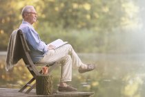Вид сбоку пожилого человека, читающего книгу на причале у озера — стоковое фото