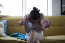Mother holding and kissing baby's belly, sitting on sofa by window — Stock Photo