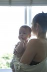 Mãe segurando pequeno bebê e olhando através da janela — Fotografia de Stock