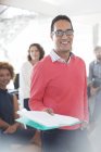 Portrait of smiling businessman wearing glasses and pink sweatshirt holding documents, office team in background — Stock Photo