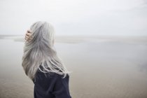 Mujer mayor con la mano en pelo largo y gris en la playa de invierno - foto de stock