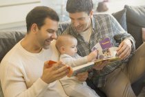 Hombre gay padres lectura libro a bebé hijo en sofá - foto de stock