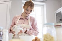 Mulher sorrindo assar, derramando açúcar em tigela na cozinha — Fotografia de Stock