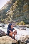 Giovane con zaino escursionismo, poggiata su rocce al fiume soleggiato — Foto stock