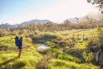 Young man with backpack hiking standing in sunny, remote field — Stock Photo
