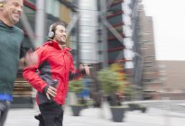 Des hommes souriants courent, portant des écouteurs sur le trottoir urbain — Photo de stock