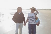 Sorridente coppia matura che si tiene per mano e cammina sulla spiaggia soleggiata — Foto stock