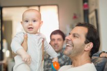 Retrato feliz macho gay padres holding bebé hijo en sala de estar - foto de stock
